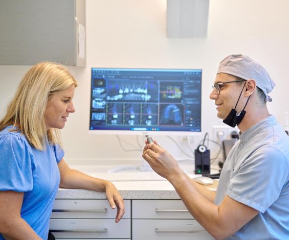 Dr. Kronauer zeigt einer Patientin ein Implantat. Im Hintergrund sieht man einen Computerbildschirm mit Röntgenaufnahmen.